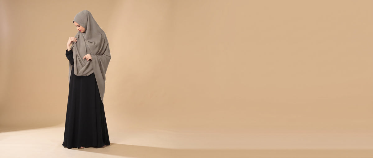 Woman wearing a grey Mehar Hijab khimar over a black abaya against a warm studio backdrop